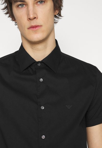 Chemise noire à manches courtes en coton avec un col classique, fermeture à boutons, et un logo ton sur ton sur la poitrine. Texture lisse.