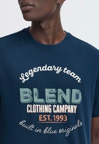 Jūras zila kokvilnas T-kreklā ar apaļu kakla izgriezumu. Iekļauj baltus un zaļus drukātus tekstus: "Leģendārā komanda BLEND Clothing Company EST. 1993."