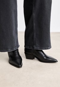 Czarne skórzane botki za kostkę z ostrym noskiem i niskim blokowym obcasem, w połączeniu z szerokimi ciemnymi dżinsami. Gładka tekstura, elegancki design.