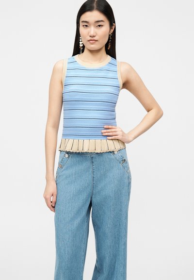 Γυναίκα που φοράει αμάνικη ριγέ πλεκτή μπλούζα σε μπλε και μπεζ με βολάν στο τελείωμα και ψηλόμεσα φαρδιά μπλε τζιν, ποζάροντας με το χέρι στη μέση.
