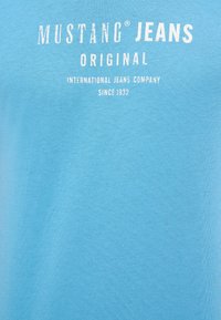 Hellblaues Baumwoll-T-Shirt mit dem weißen Schriftzug "MUSTANG JEANS", ausgestattet mit einer glatten Textur und einem Rundhalsausschnitt.