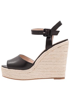 Sandale compensée en cuir noir avec un bout ouvert, une bride de cheville réglable et une semelle intermédiaire en corde de jute texturée. Fini lisse et forme arrondie.