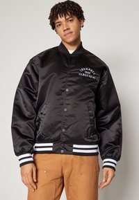Chaqueta bomber de satén negra con texto gráfico blanco en el pecho, puños de canalé a rayas y cierre de botones a presión. Llevada con pantalones beige.