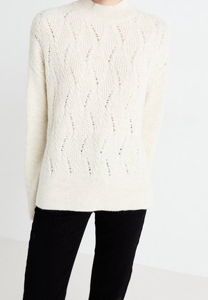 Pull en maille couleur crème avec des détails de motif en vague, porté avec un pantalon noir, présenté des épaules aux hanches sur un fond uni.