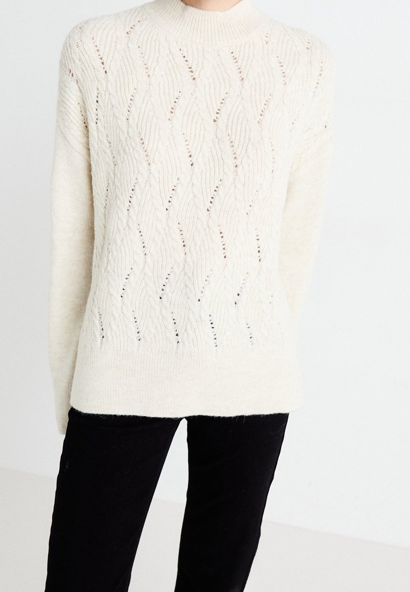 Pull en maille couleur crème avec des détails de motif en vague, porté avec un pantalon noir, présenté des épaules aux hanches sur un fond uni.