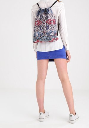 Osoba ubrana w białą koszulę, niebieską spódnicę, białe trampki i kolorowy plecak ze ściągaczem, odwrócona tyłem na jednolitym tle.