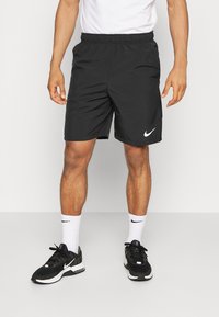 Svarta Nike-shorts med elastisk midja och vitt logotyp, i kombination med svarta sneakers och vita strumpor, stående mot en neutral bakgrund.