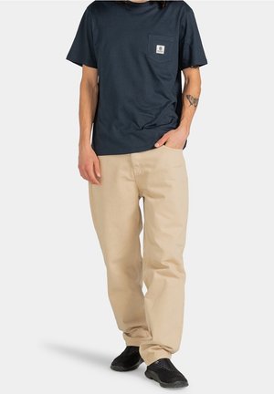 Homme portant une chemise bleu marine à manches courtes avec poche, pantalon beige, chaussures noires et un tatouage de papillon sur l'avant-bras gauche, main dans la poche.
