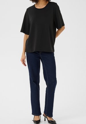 Nainen, jolla on yllään väljät mustat lyhythihainen paita, tummansiniset suorat farkut ja mustat korkokengät, joissa on hihna kantapään takana.