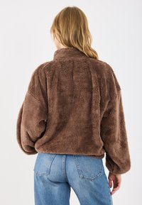 Brązowa, puszysta kurtka typu cropped z wysokim kołnierzem i oversize'owymi rękawami, w połączeniu z niebieskimi dżinsami z wysokim stanem. Widoczna miękka tekstura.