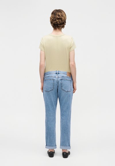 Άτομο με κοντά καστανά μαλλιά, φορεμένο μπεζ κοντομάνικο πουκάμισο, ανοιχτό μπλε καμπαρντινέ παντελόνια που είναι σηκωμένα και μαύρα παπούτσια, στέκεται γυρισμένο με την πλάτη.