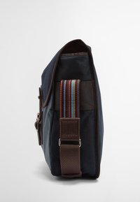 Saco de lona azul escuro com um detalhe em couro castanho e uma alça de ombro riscada. Apresenta um fecho em aba e detalhes em metal prateado.