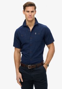 Camisa de manga corta en tejido texturizado color marino, con dos bolsillos en el pecho, botones marrones y un collar doblado. Se lleva con pantalones oscuros y un reloj.