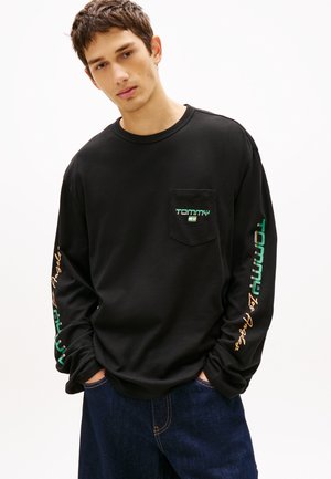 Jauns vīrietis, kas valkā melnu garās piedurknes krekli ar mazu kabatas logotipu un krāsainiem uzrakstiem uz piedurknēm, savienots ar vaļīgām tumši zilām džinsām.