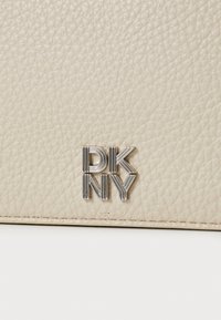 Kreemja tekstuuriga nahast kott, millel on hõbedane DKNY logo. Pind on pehme tekstuuriga, rõhutades sügavust ja mõõdet.