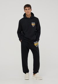 Felpa nera con tasca frontale e stampa grafica arancione. Abbinata a pantaloni jogger neri con stampa simile. Materiale misto cotone.