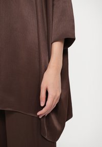 Καφέ, υφασμάτινη μπλούζα με υφή, ευρύ λαιμόκοψη και χαλαρή γραμμή. Το ύφασμα πέφτει απαλά, με μια διακριτική λάμψη και κομψή αίσθηση.