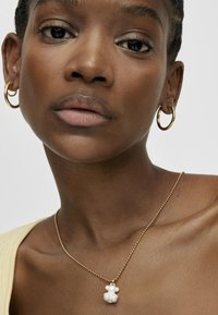 Collier en chaîne dorée avec un pendentif en forme d'ours blanc. Le pendentif a une texture brillante. Le modèle porte des boucles d'oreilles créoles et un haut jaune.