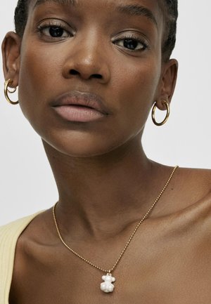 Goldene Kette mit einem weißen Bärenanhänger. Der Anhänger hat eine glänzende Oberfläche. Das Modell trägt Creolen und ein gelbes Oberteil.