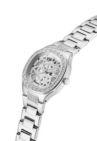 Montre squelette argentée avec un cadran transparent, ornée de cristaux transparents sur la lunette et le bracelet. Acier inoxydable, forme ovale, finition polie.