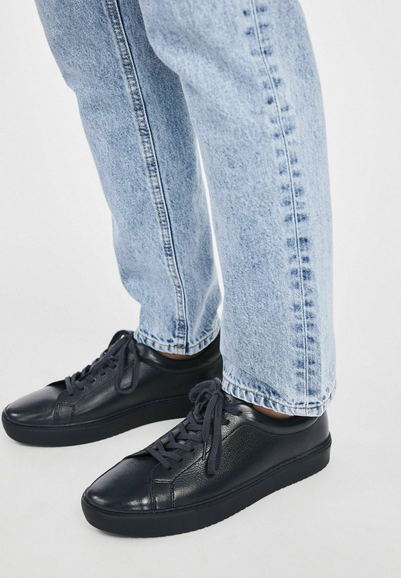Czarne skórzane trampki o gładkiej strukturze, okrągłym nosku i płaskiej podeszwie, zestawione z jasnoniebieskimi dżinsami o prostym kroju.