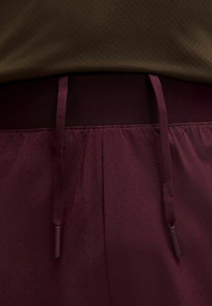 Nahaufnahme von dunkelroten Sportshorts mit Kordelzug und kleinen perforierten Löchern, kombiniert mit einem braunen, strukturierten Hemd.