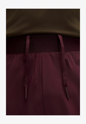 Nahaufnahme von dunkelroten Sportshorts mit Kordelzug und kleinen perforierten Löchern, kombiniert mit einem braunen, strukturierten Hemd.