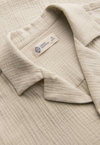 Col de chemise en tissu beige texturé plié avec une étiquette visible indiquant Ombre Heritage, taille moyenne, 48 EUR.