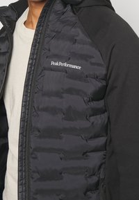 Sort jakke med en quiltet, puffy design på den ene side og glat stof på den anden, med lynlås lukning og "PeakPerformance" branding.