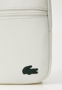 Borsa a pochette bianca con superficie testurizzata, caratterizzata da un logo di coccodrillo verde e due scomparti con zip lungo la parte superiore.