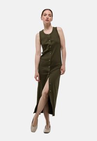 Robe midi sans manches vert olive avec un devant à boutons, une fente sur le côté et une texture côtelée. Portée avec des chaussures de couleur claire.
