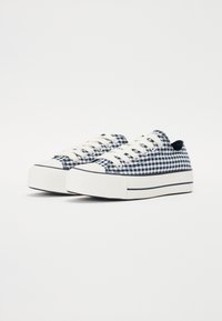 Une paire de baskets basses avec un motif vichy bleu et blanc, une épaisse semelle en caoutchouc blanche et des lacets blancs sur un fond blanc uni.