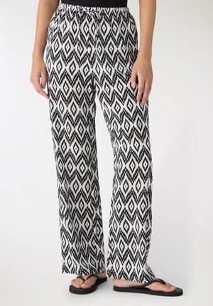 Donna che indossa pantaloni larghi a motivo di diamanti bianco e nero con coulisse in vita e infradito nere, in piedi davanti a uno sfondo bianco.