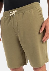 Olive green shorts made of soft cotton blend, featuring an elastic waistband with drawstring and side pockets, with logo detail on the leg.