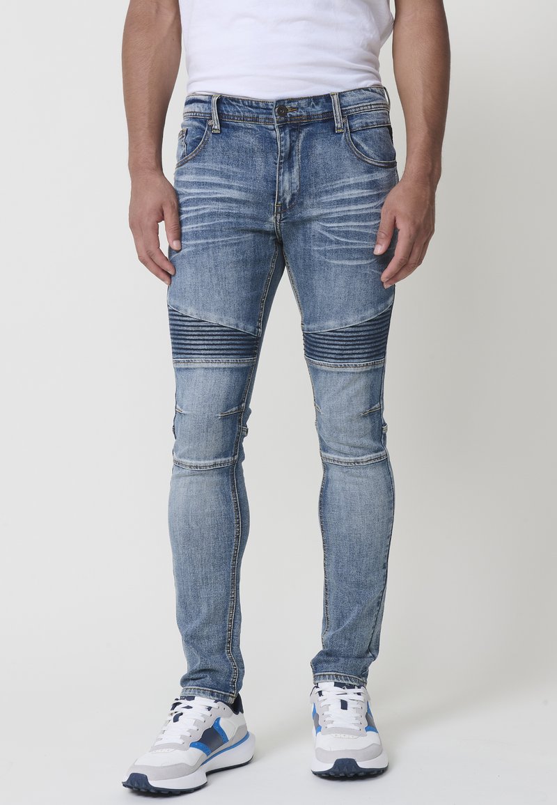 Jeans de denim ajustados en azul con paneles de rodilla acanalados, detalles desgastados y diseño de cinco bolsillos. Combinados con zapatillas deportivas blancas y azules.