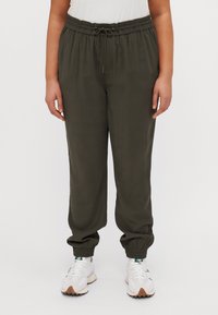 Pantalones jogger en verde oliva, ligeros, con cinturilla elástica y cordón. Cuentan con puños elásticos y un corte relajado. Combinados con zapatillas blancas.