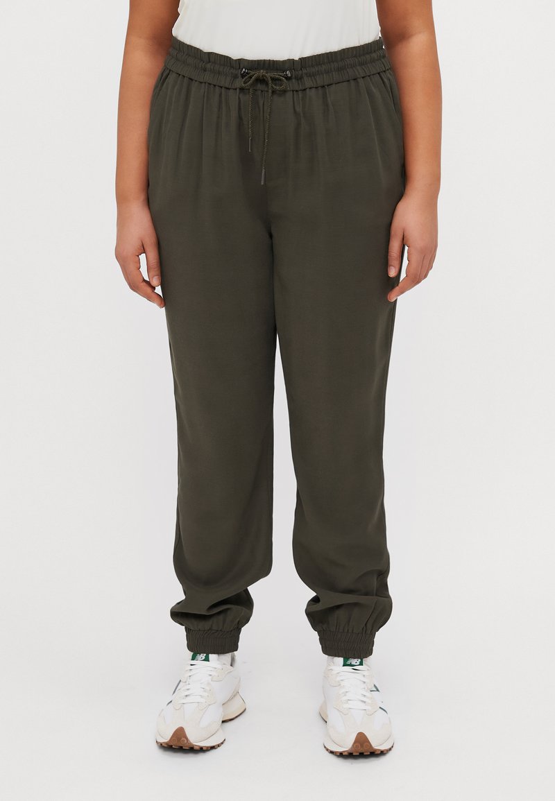 Pantalones jogger en verde oliva, ligeros, con cinturilla elástica y cordón. Cuentan con puños elásticos y un corte relajado. Combinados con zapatillas blancas.