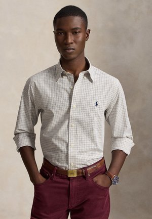 Jeune homme portant une chemise légère à carreaux avec manches retroussées, pantalon bordeaux, ceinture en cuir marron et montre à cadran bleu.