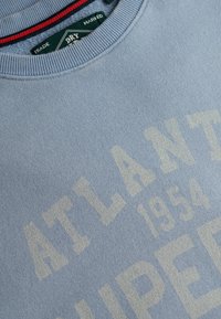 Sweat-shirt bleu clair à col rond avec texte crème pâle "ATLANTA 1954" et étiquette de marque verte à l'intérieur du col.
