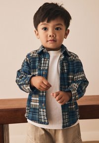 Skjorte med tern i blå, grønne og brune nuancer, med knapper, lagdelt over en hvid t-shirt, båret med beige shorts. Glat stoftekstur.