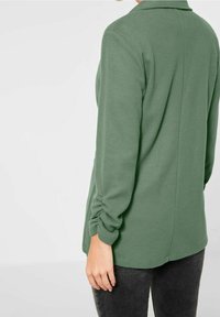 Zelený top s dlouhým rukávem a volným střihem, s límečkem a našitými manžetami, vyrobený z měkké látky, v jednobarevném provedení bez vzorů.