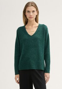 Pulover verde tricotat cu decolteu în V, tiv franjurat și mâneci lungi. Textură moale și tăietură lejeră, purtat peste pantaloni închiși la culoare.