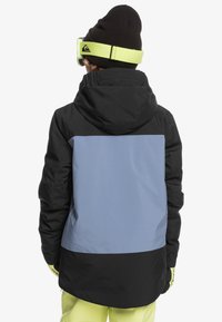 Giacca da sci nera e azzurra con cappuccio. Presenta un design color-block e un tessuto testurizzato. Indossata con un berretto nero e occhiali da sci verdi.