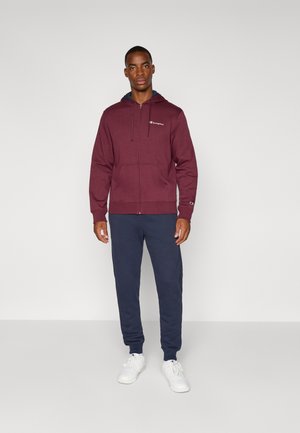 Felpa con zip color marrone bordeaux con interno blu, caratterizzata da un logo frontale, abbinata a pantaloni della tuta blu navy e sneakers bianche. Tessuto liscio.