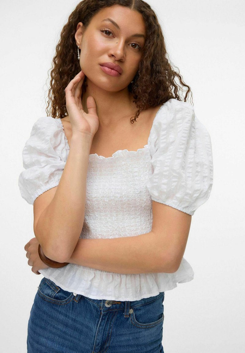 Blusa bianca con maniche a sbuffo e scollatura quadrata, corpino testurizzato e orlo smerlato. Abbinata a jeans in denim a vita alta.