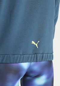 Blå sweatshirt med ribbad nederkant och gul Puma-logotyp, kombinerad med blå och lila mönstrade leggings med en slät och glänsande yta.