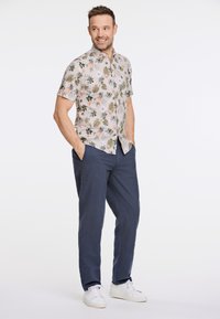 Camicia a maniche corte, di colore chiaro, con un motivo floreale verde e rosa, abbinata a pantaloni larghi di colore blu scuro e sneakers bianche.