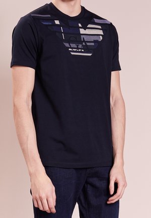 Homme portant un t-shirt noir à manches courtes avec un grand logo d'aigle stylisé en nuances de bleu et de gris sur la poitrine, associé à un jean foncé.