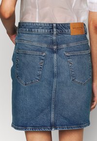 Žena v modré denimové sukni se dvěma zadními kapsami a koženou etiketou "SELECTED FEMME", spárované s polotransparentním bílým topem.