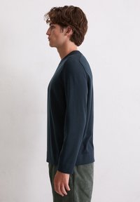 Chemise à manches longues bleu marine avec un col rond et des poignets côtelés. Portée avec un pantalon en velours côtelé vert, mettant en valeur une texture lisse.